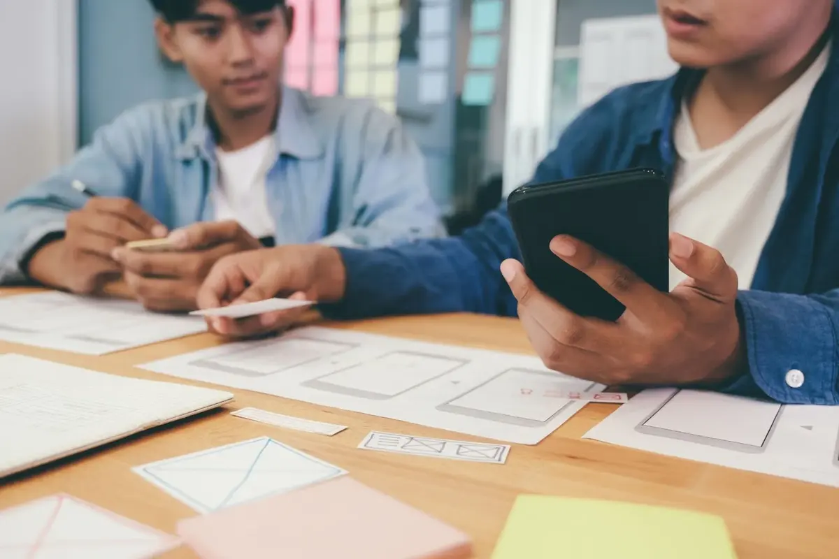 "Dos personas trabajando en una mesa con hojas impresas que representan pantallas de teléfono celular. Una de las personas sostiene un teléfono celular en la mano mientras coloca un papel sobre estas pantallas impresas."